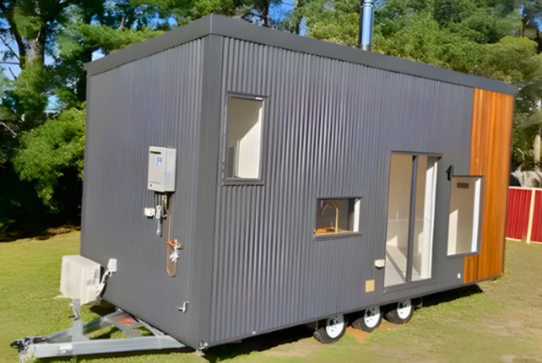 Modern tiny house on wheels with gray corrugated metal exterior and orange/red accent, featuring multiple windows and door, parked on grass with trees in background