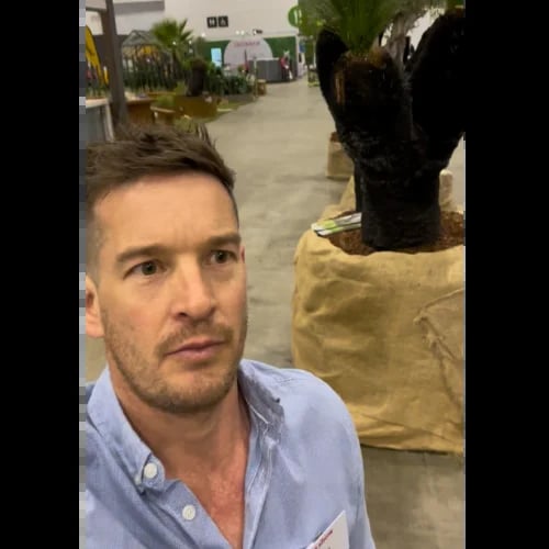 Man in light blue shirt taking selfie indoors with potted plant and garden center background visible